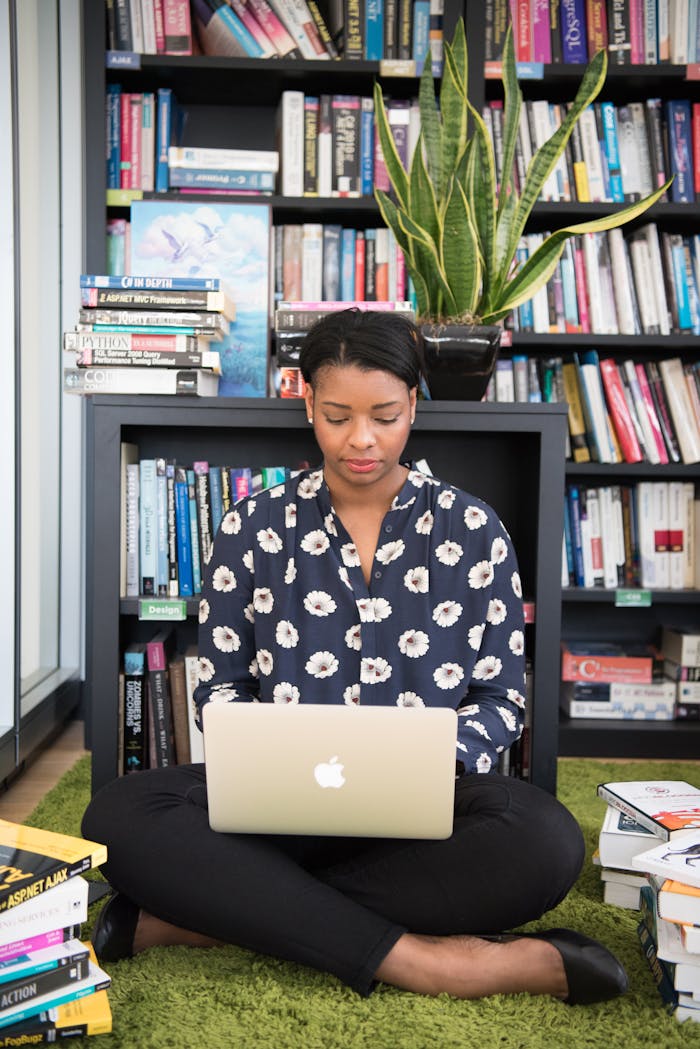 Woman sitting on carpet with laptop, surrounded by bookshelves and plants, focused on study.